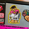 A cartoon-style die-cut sticker of a figure drawing in a notebook, featuring a pink shirt, brown skin tone, and a yellow border, shown on a glittery grey surface next to other stickers.