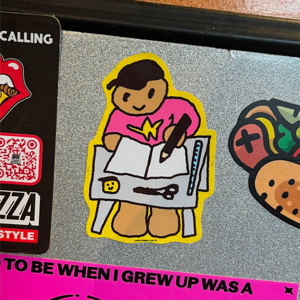 A cartoon-style die-cut sticker of a figure drawing in a notebook, featuring a pink shirt, brown skin tone, and a yellow border, shown on a glittery grey surface next to other stickers.