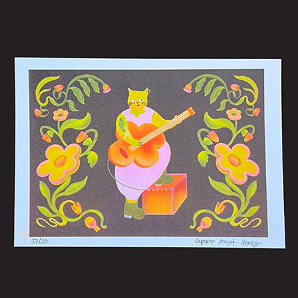 A colourful art print featuring a stylised green cat playing an orange guitar on a dark grey background, framed by yellow and pink flowers with green vines. The print has a white border and is photographed against a solid black background.