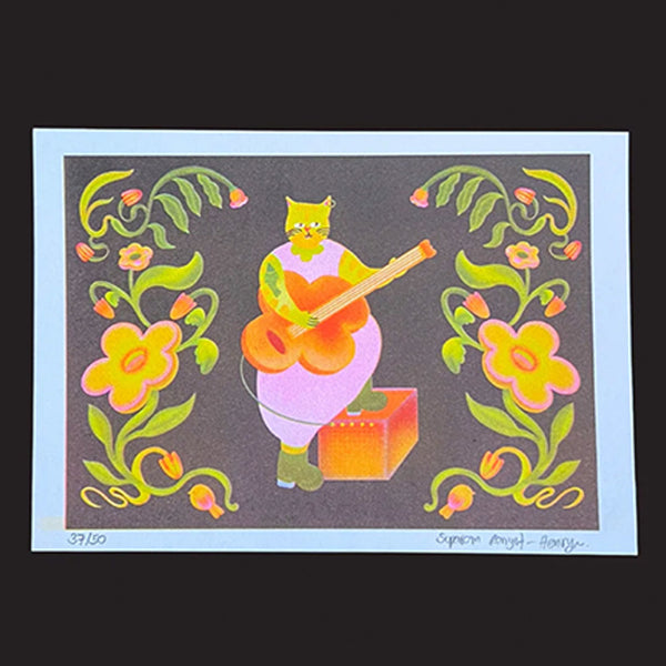 A colourful art print featuring a stylised green cat playing an orange guitar on a dark grey background, framed by yellow and pink flowers with green vines. The print has a white border and is photographed against a solid black background.