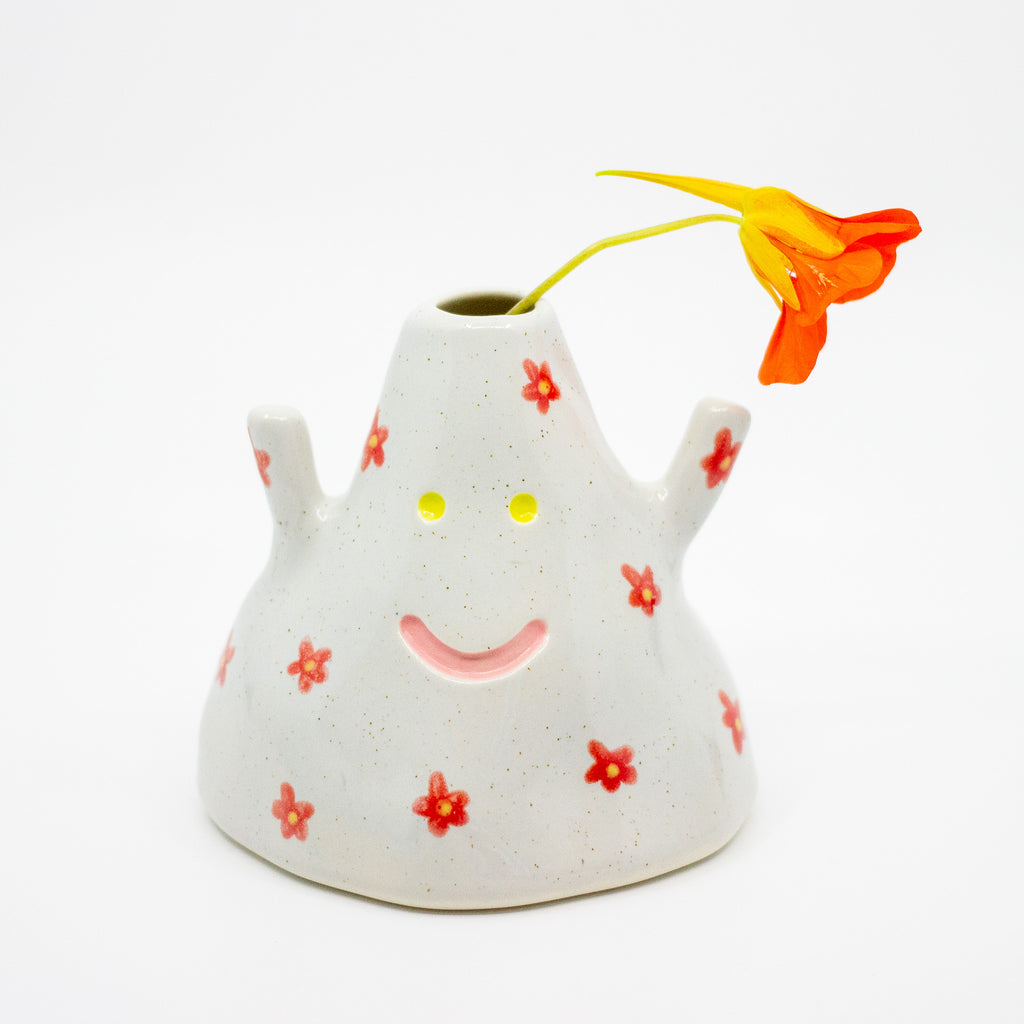 Ceramic vase with a smiley face design and a single orange flower on a white background