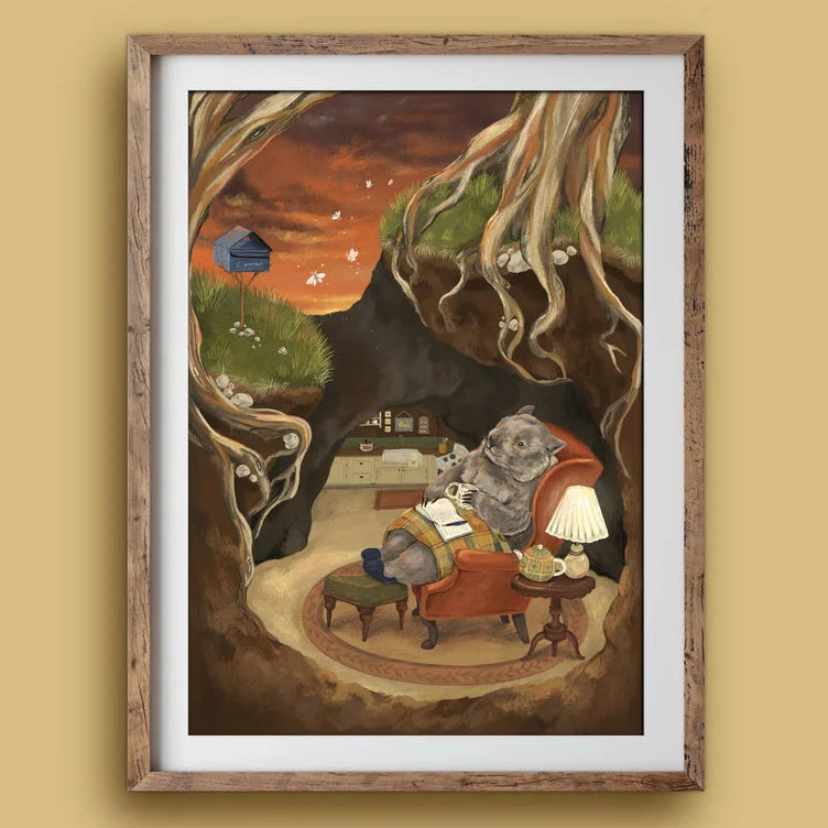 A framed art print featuring a whimsical illustration of a wombat lounging in an orange armchair inside a tree hollow, set against a warm beige background.