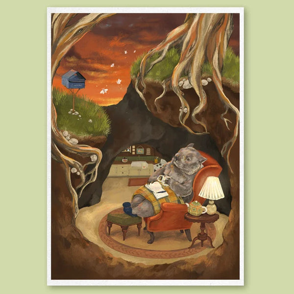 A framed art print featuring a whimsical illustration of a wombat sitting in an armchair inside a tree hollow, with a small house under a tree and an orange sky in the background, displayed on a light green background.