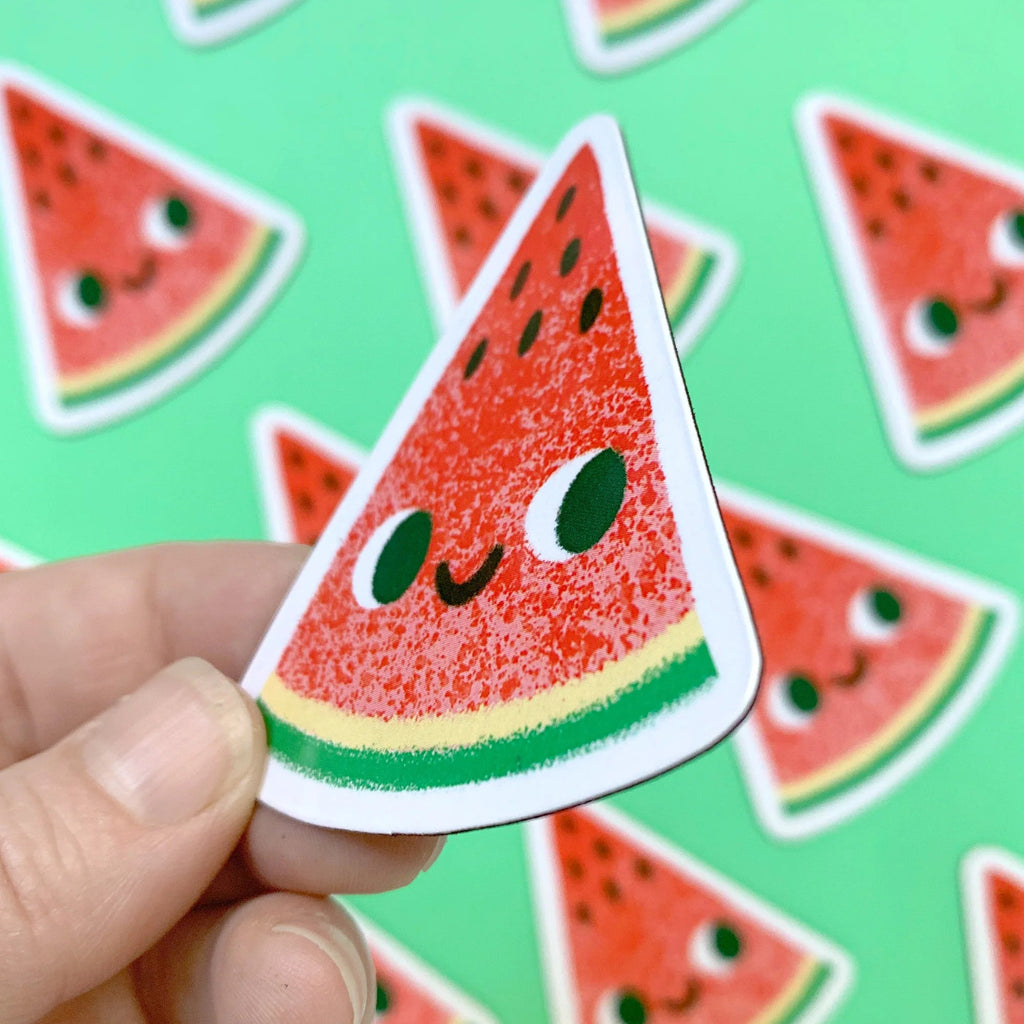 Hand holding a cute watermelon slice magnet with more stickers in the background on a green surface.