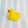 A small glossy yellow duck ceramic chopstick holder with a red beak and a black eye, standing on a light blue and white striped background.