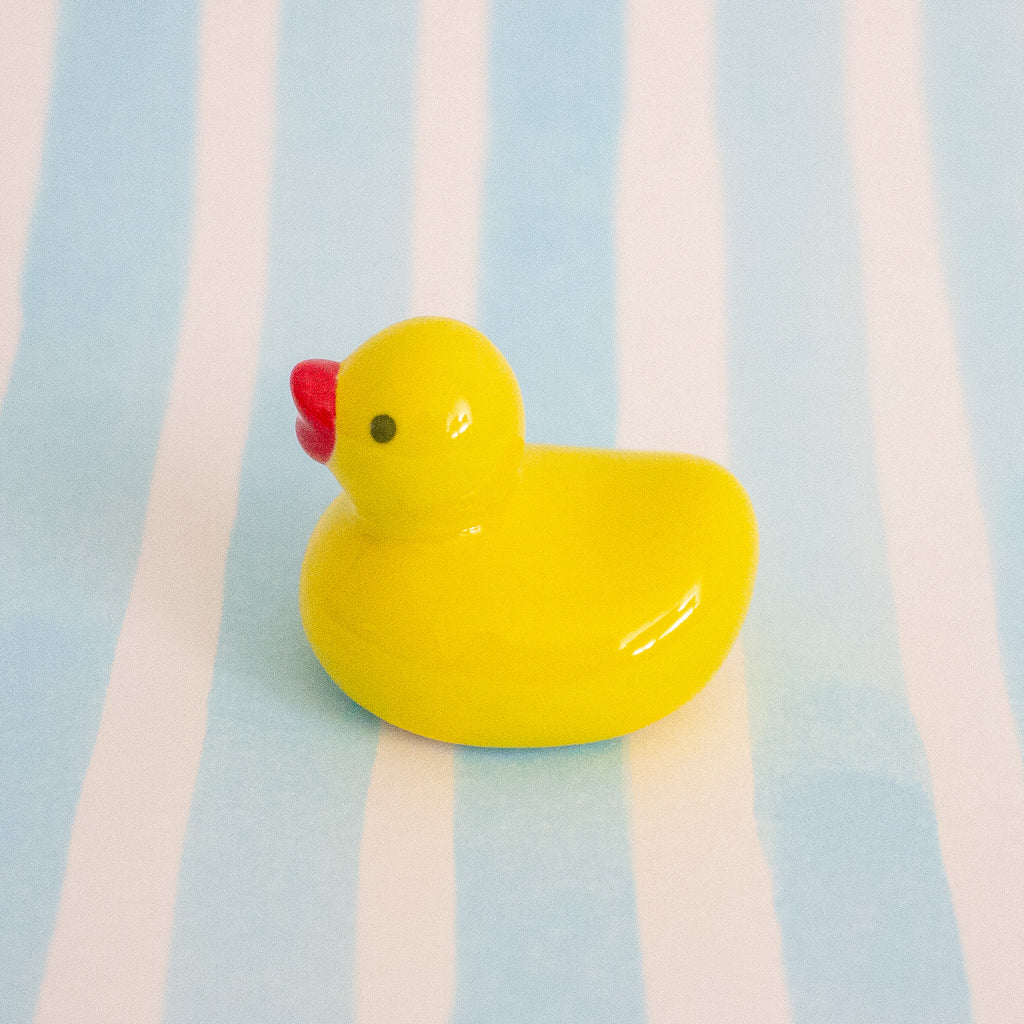 A small glossy yellow duck ceramic chopstick holder with a red beak and a black eye, standing on a light blue and white striped background.