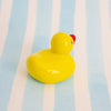 A small glossy yellow duck figurine with a red beak sits on a light blue and white striped background.