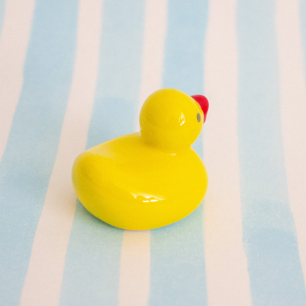 A small glossy yellow duck figurine with a red beak sits on a light blue and white striped background.