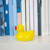 A small glossy yellow rubber duck with a red beak sits on a white shelf in front of a row of colourful books.