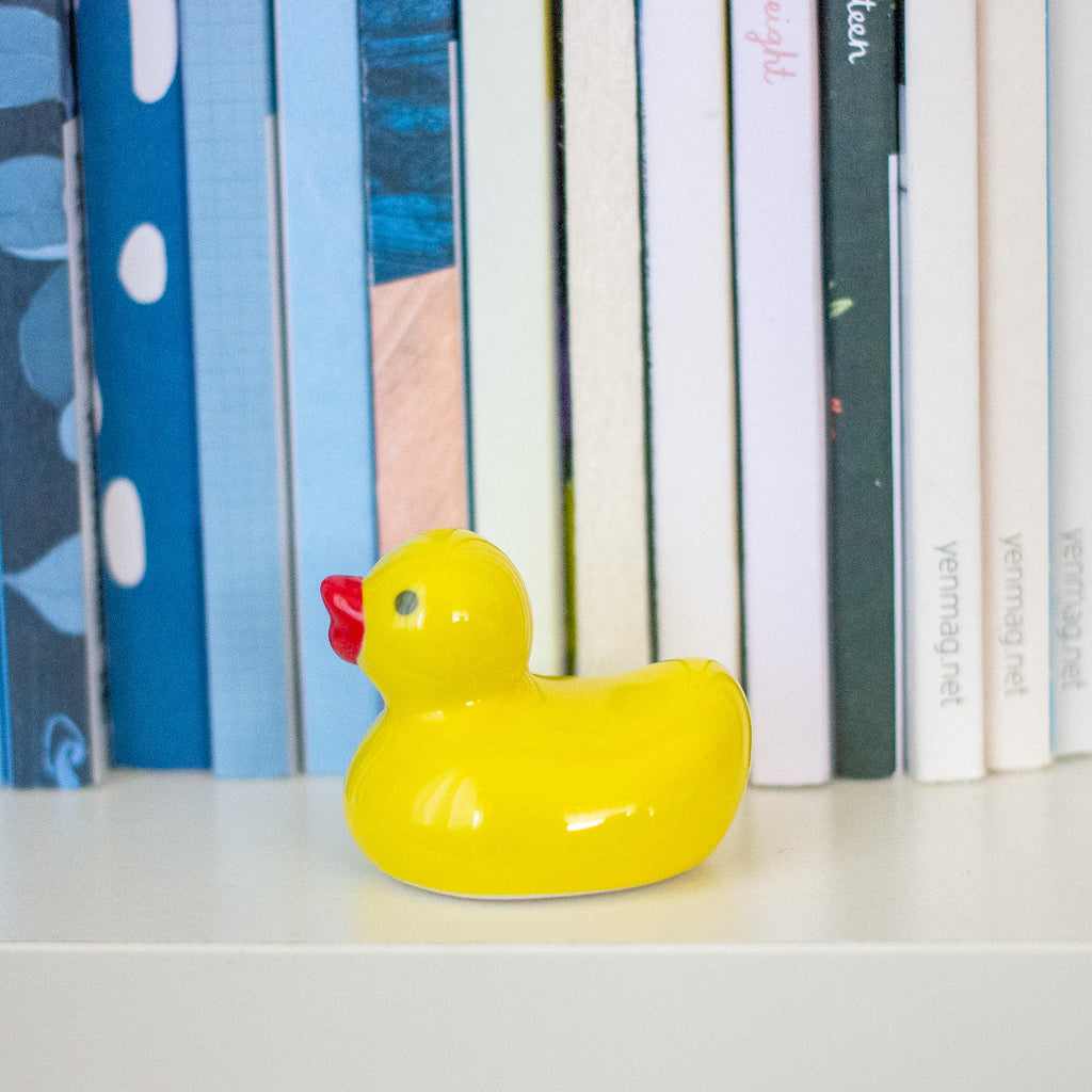 A small glossy yellow rubber duck with a red beak sits on a white shelf in front of a row of colourful books.