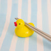 A small glossy yellow duck-shaped chopstick holder with a red beak, holding a pair of metal chopsticks, set against a light blue and white striped background.