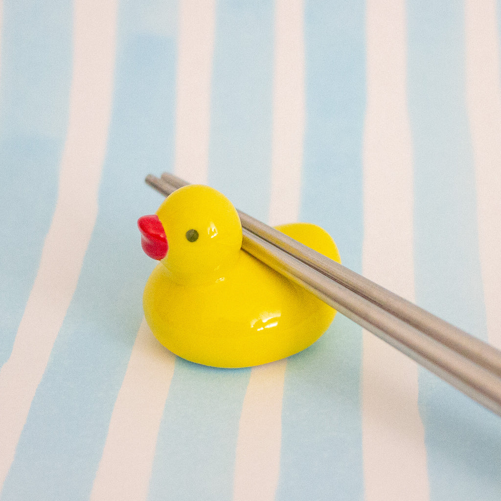 A small glossy yellow duck-shaped chopstick holder with a red beak, holding a pair of metal chopsticks, set against a light blue and white striped background.