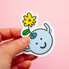 A hand holding a die-cut magnet featuring a light blue smiling character holding a yellow flower with a green leaf, set against a solid pink background.