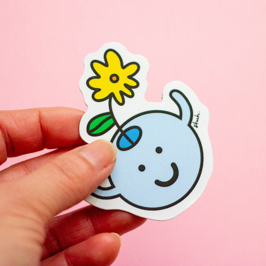 A hand holding a die-cut magnet featuring a light blue smiling character holding a yellow flower with a green leaf, set against a solid pink background.