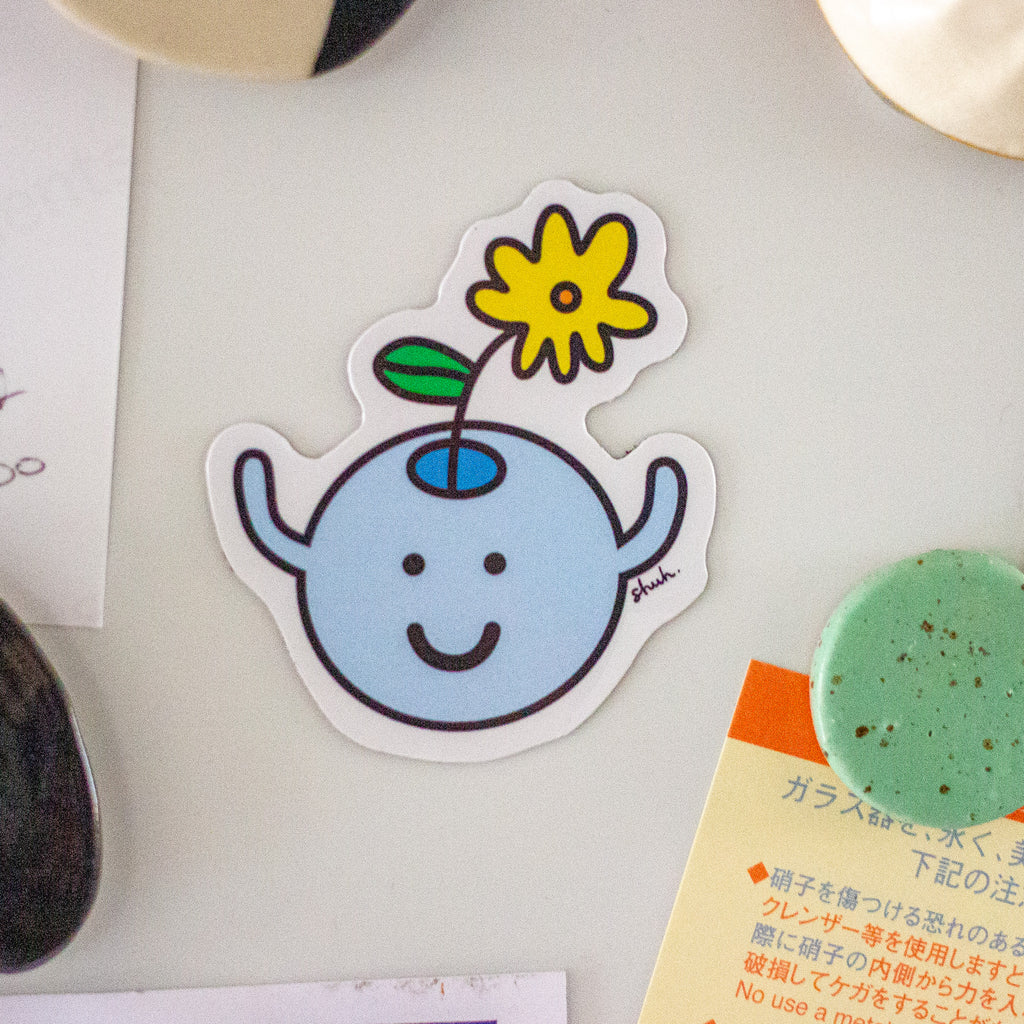 A die-cut magnet featuring a cute light blue character with a smiling face and a yellow flower on its head, placed on a light surface with partial surrounding objects visible.
