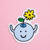 A die-cut magnet featuring a cute cartoon character with a light blue round head, black smile, simple face, and a yellow flower with a green leaf on its head, shown on a solid pink background.