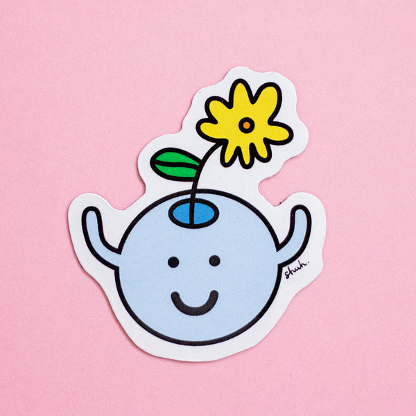 A die-cut magnet featuring a cute cartoon character with a light blue round head, black smile, simple face, and a yellow flower with a green leaf on its head, shown on a solid pink background.