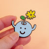 A small enamel pin shaped like a smiling light blue character with a yellow flower on its head, held between fingers against a soft peach background.