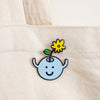 A small enamel pin shaped like a light blue smiling face with a yellow flower and green leaf on a stem, attached to a cream canvas tote bag.
