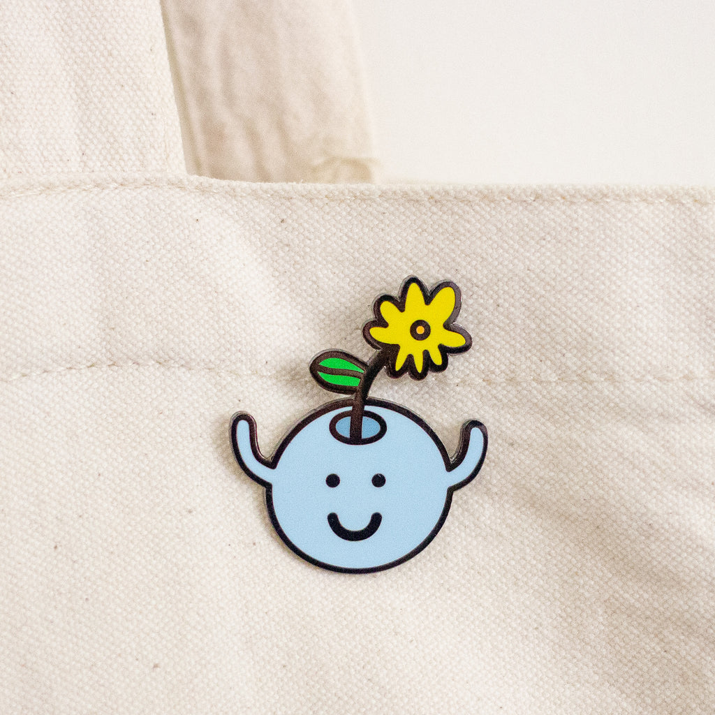 A small enamel pin shaped like a light blue smiling face with a yellow flower and green leaf on a stem, attached to a cream canvas tote bag.