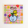 A cute enamel pin featuring a light blue round character with a smiling face and a yellow flower on its head, displayed on a colourful pink and beige plaid backing card with the brand name 'shuh.'.