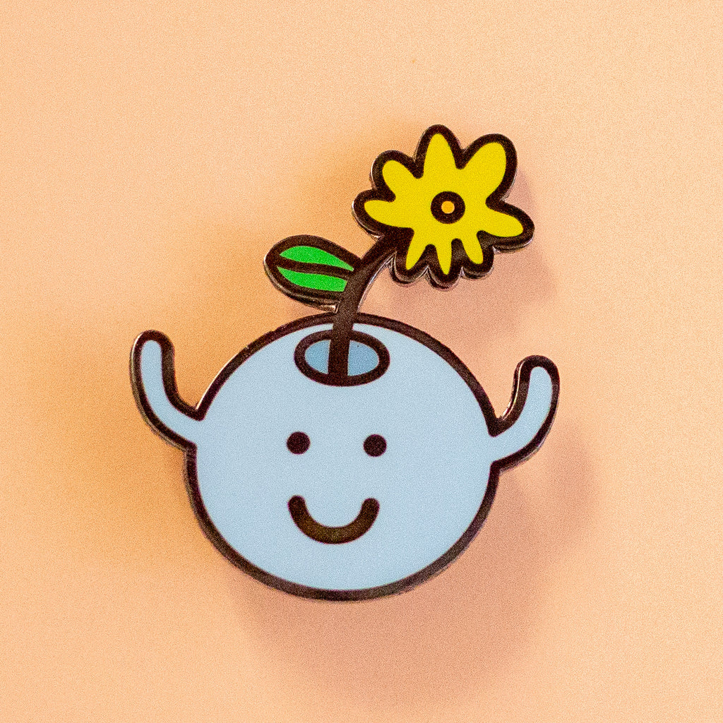 A cute enamel pin featuring a light blue round character with a smiling face, small arms, and a yellow flower growing from its head, set against a solid peach background.