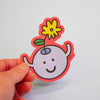 A hand holding a die-cut sticker featuring a cute cartoon character with a yellow flower on its head, set against a plain light grey background.