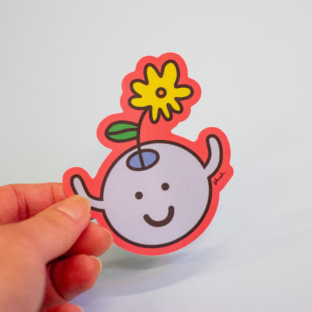A hand holding a die-cut sticker featuring a cute cartoon character with a yellow flower on its head, set against a plain light grey background.