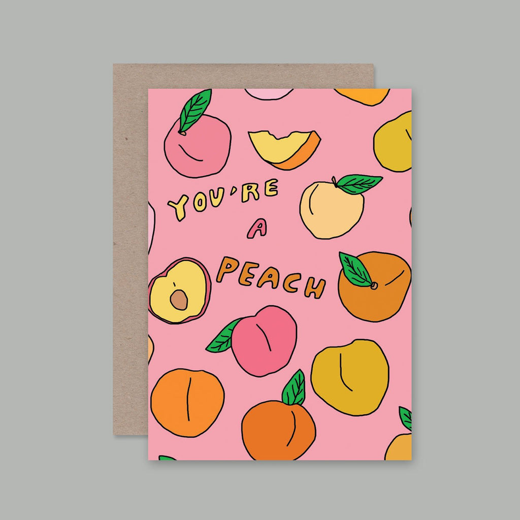 A pink greeting card with a peach and fruit illustration pattern and the text 'YOU'RE A PEACH', shown with a brown kraft envelope on a plain grey background.