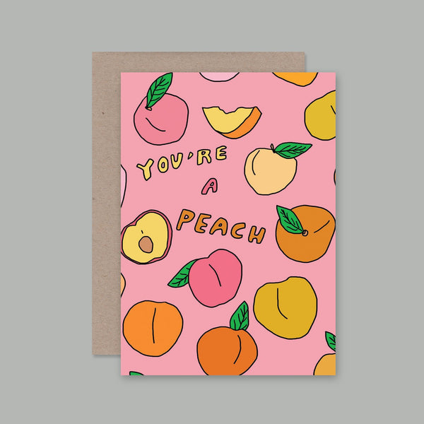 A pink greeting card with a peach and fruit illustration pattern and the text 'YOU'RE A PEACH', shown with a brown kraft envelope on a plain grey background.