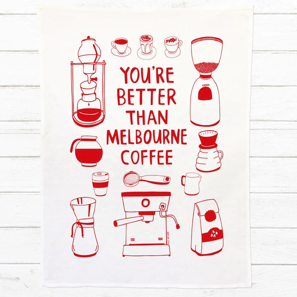 A white kitchen towel featuring red line drawings of coffee-related items and the text 'YOU'RE BETTER THAN MELBOURNE COFFEE' in red, displayed flat against a white wood plank background.