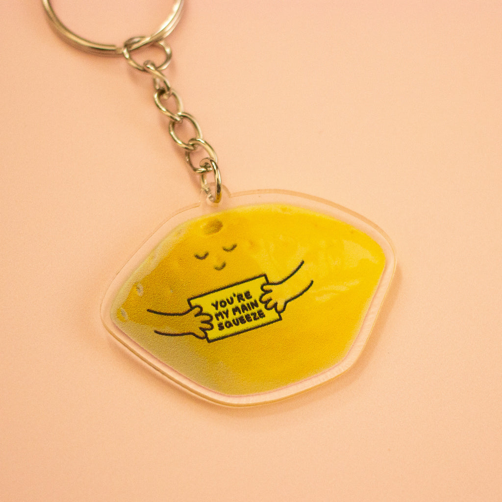 A keychain featuring a yellow lemon-shaped charm with a cute sleeping face and the text 'YOU'RE MY MAIN SQUEEZE' in black, attached to a silver metal keyring, set against a peach background.