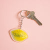 A keychain featuring a yellow lemon-shaped charm with a cute sleeping face and the text 'YOU'RE MY MAIN SQUEEZE' in black, attached to a silver metal keyring and key, set against a peach background.