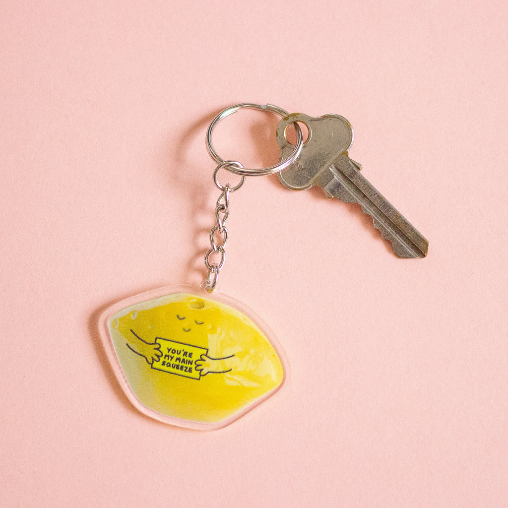 A keychain featuring a yellow lemon-shaped charm with a cute sleeping face and the text 'YOU'RE MY MAIN SQUEEZE' in black, attached to a silver metal keyring and key, set against a peach background.