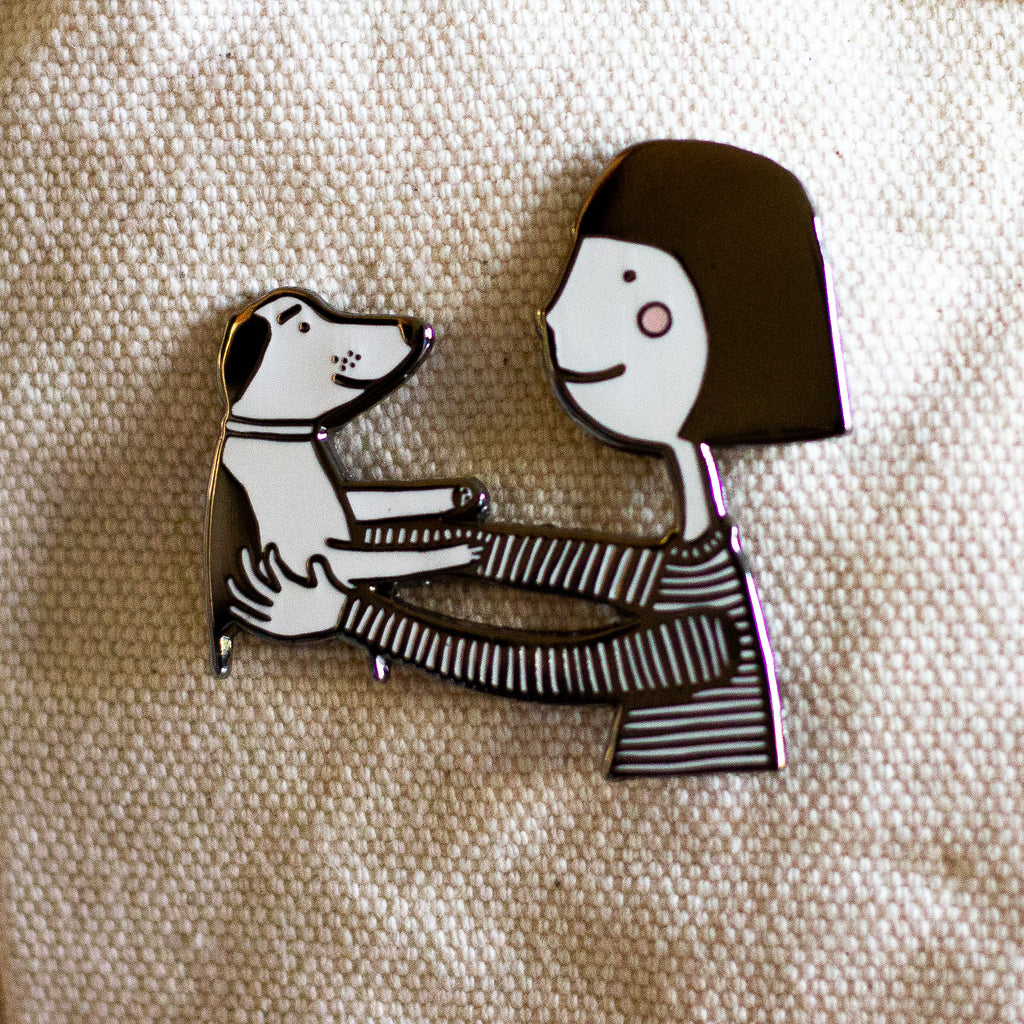 The pin features a girl holding a dog. Girl has pink cheeks. 