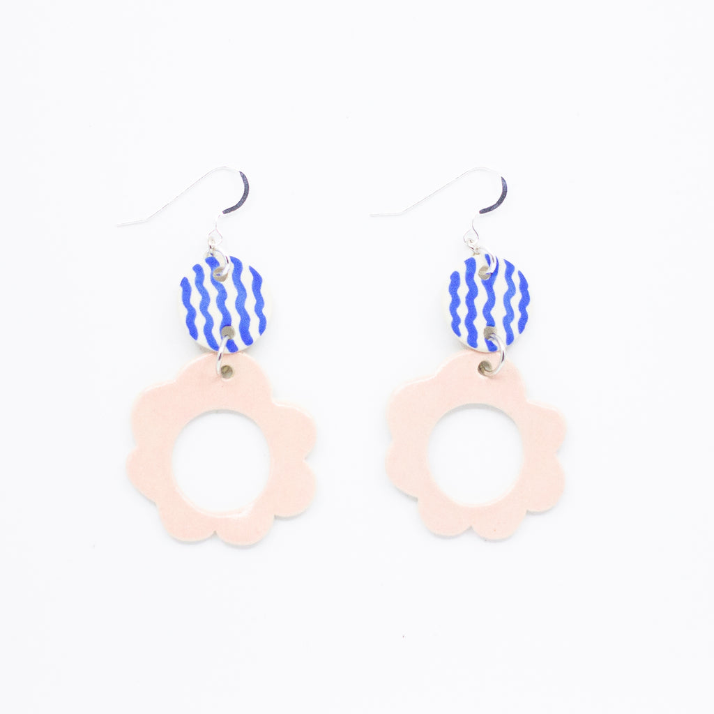 Two drop earrings with sterling silver hooks. Each earring has two ceramic pendants, the first is circular with wavy blue handpainted lines. The second is a larger circular pendant with pink colour and flower shape. On white background. 