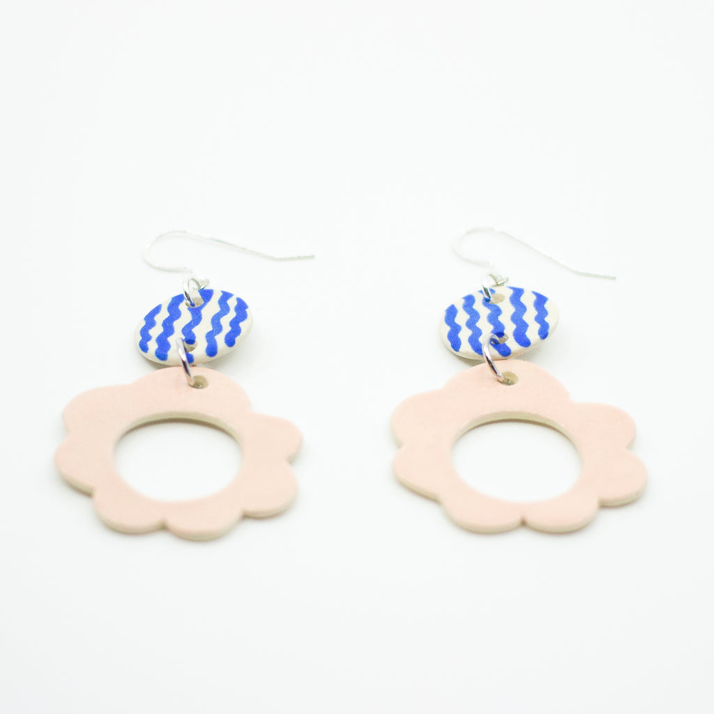 A flat angle of two drop earrings with sterling silver hooks. Each earring has two ceramic pendants, the first is circular with wavy blue handpainted lines. The second is a larger circular pendant with pink colour and flower shape. 