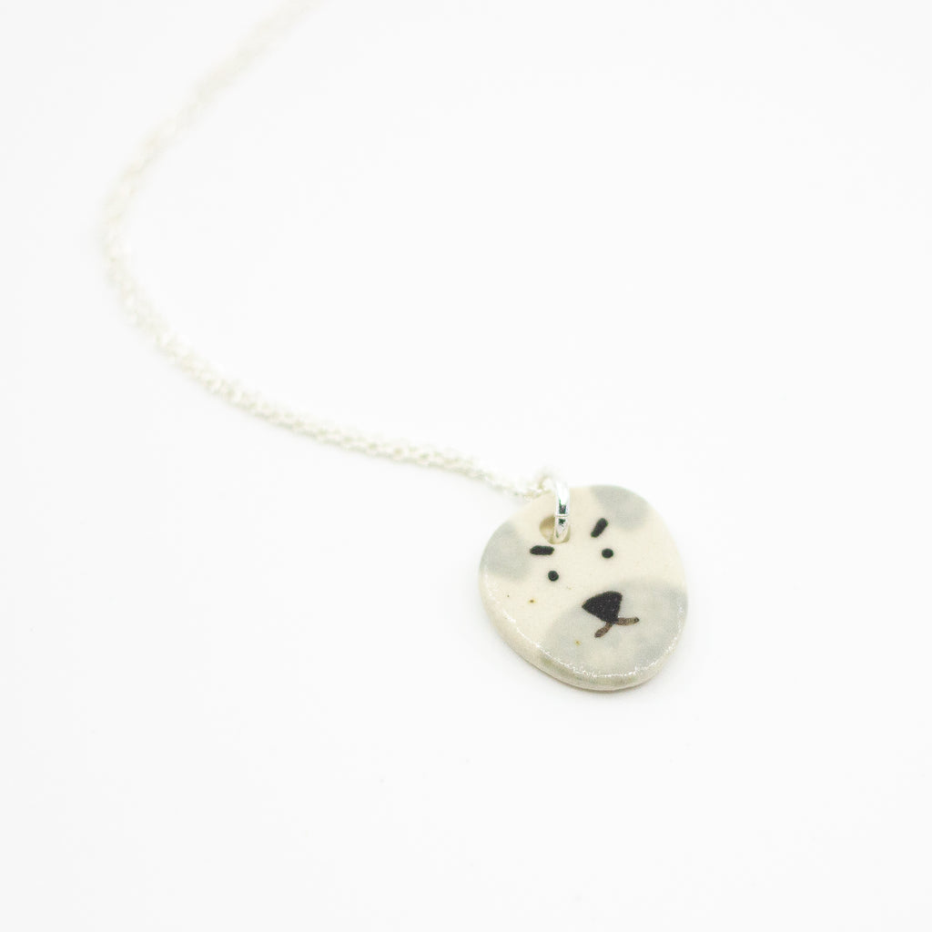 Sterling silver necklace featuring a round ceramic pendant hand-painted with a light grey and cream dog face, including expressive eyebrows and a small brown mouth, displayed on a white background.