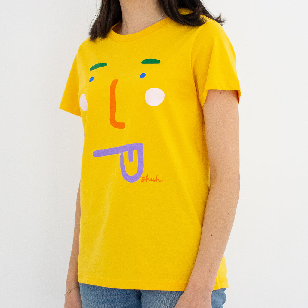 A model wearing the boo tee. Mustard coloured tee, with a stylised face with tongue poking out. 
