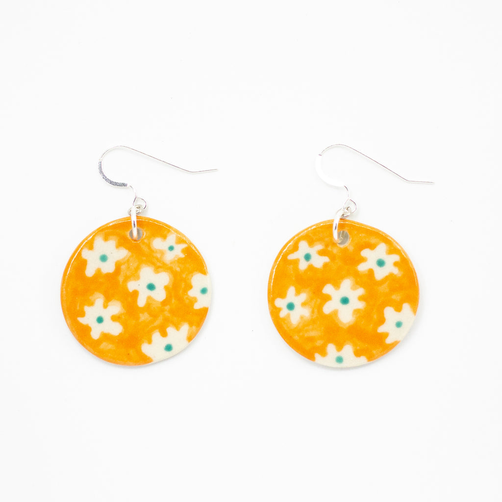A pair of handmade ceramic earrings with round orange drops, decorated with a pattern of white flowers and green centres. Finished with silver hooks for pierced ears.