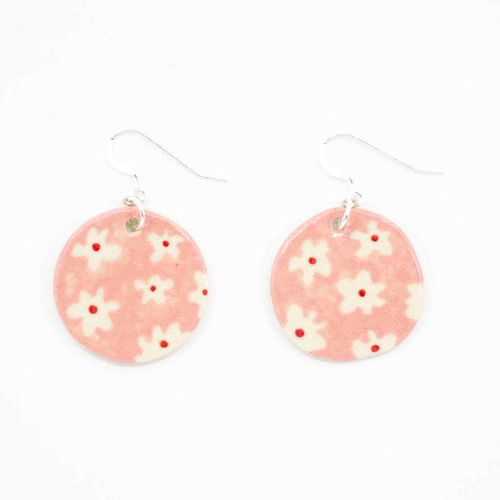 A pair of handmade ceramic earrings with round soft pink drops, decorated with a pattern of white flowers and red centres. Finished with silver hooks for pierced ears.
