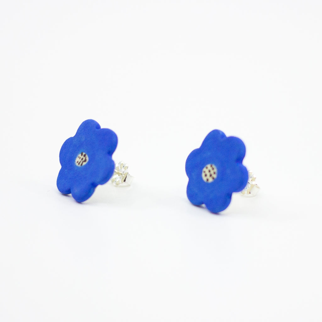 Side view of a pair of handmade ceramic stud earrings shaped like bright blue flowers with speckled cream and black centres. Finished with sterling silver stud backings for pierced ears.
