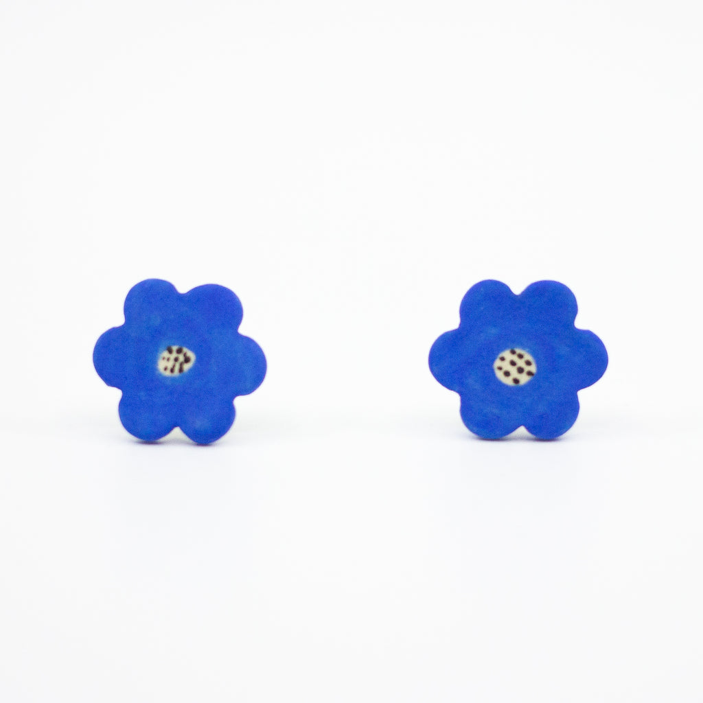 A pair of handmade ceramic stud earrings shaped like bright blue flowers with speckled cream and black centres. Finished with sterling silver stud backings for pierced ears.