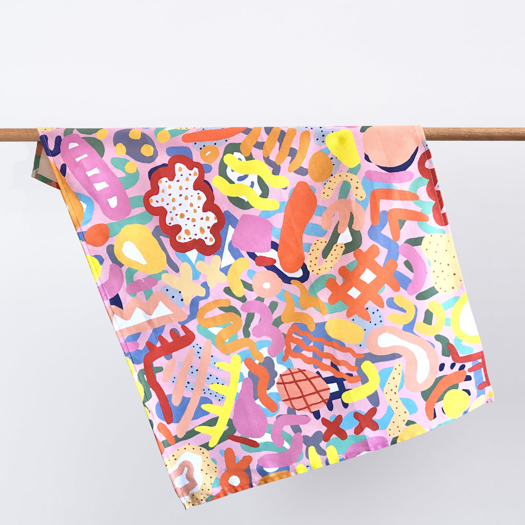 Coral cotton tea towel, hanging from a wooden rack. Print is bright, multicoloured, and is about the microscopic sea life in a coral reef. 