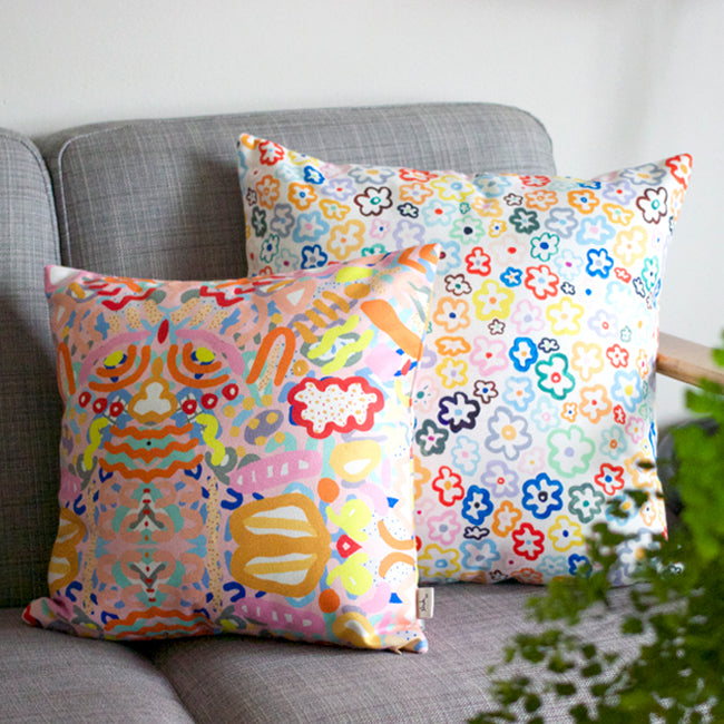 Front view of the daisy cushion cover, alongside the smaller Coral cushion cover. With illustrated multi-coloured daisies on a white background. Sitting on a couch. 