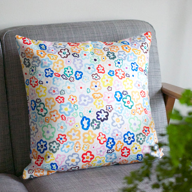 Front view of the daisy cushion cover. With illustrated multi-coloured daisies on a white background. Sitting on a couch. 