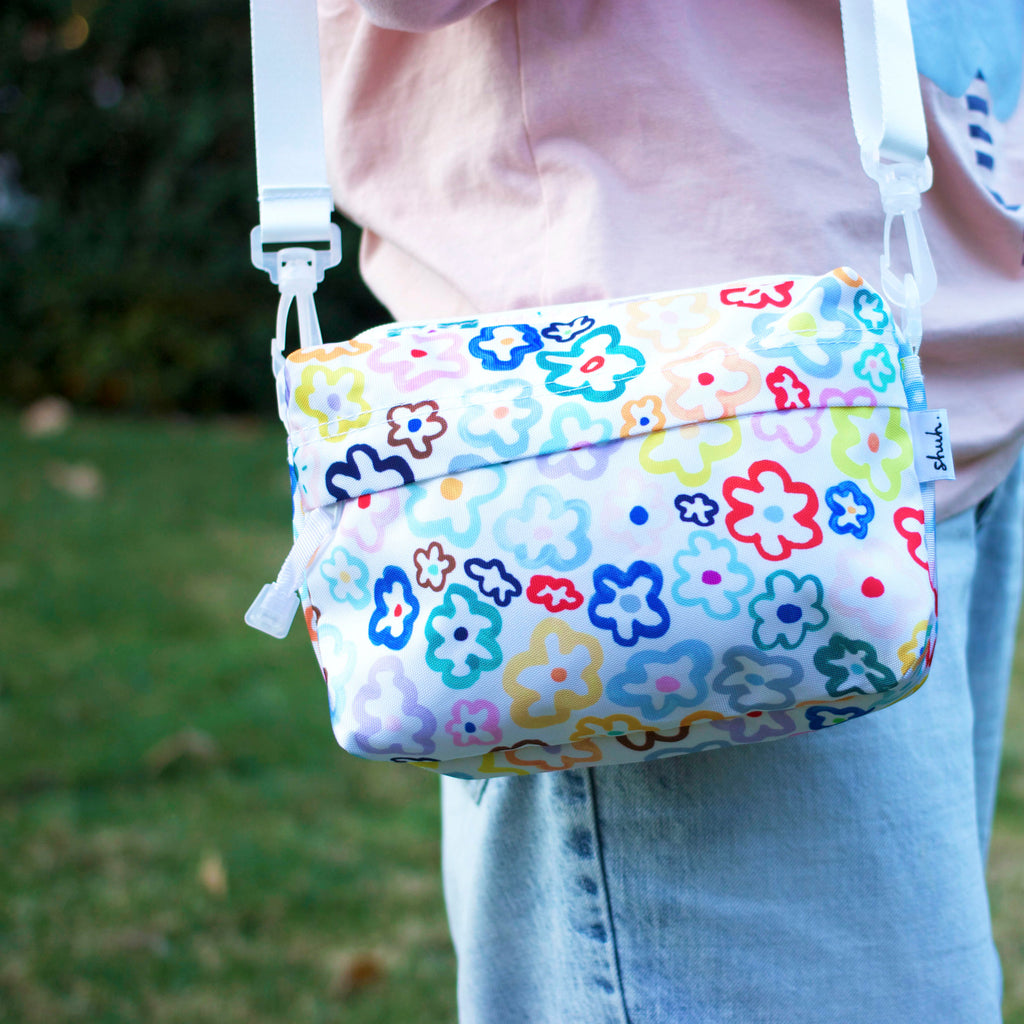 A picture of the daisy everyday bag, carried on a persons shoulder. Multicoloured daisy illustrations on a white background. 