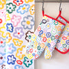 The Daisy print on a tea towel, oven mitt, and pot holder. Multi-coloured daisies on a white background