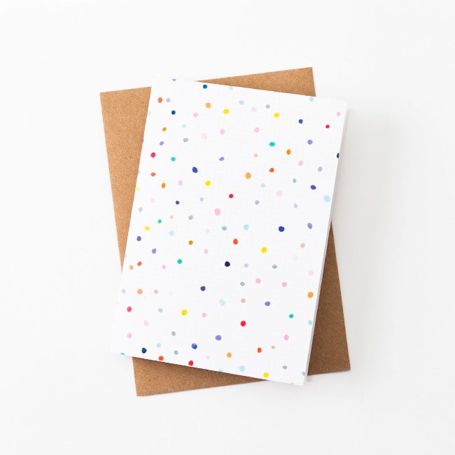 A front view of the Day Pop greeting card with recycled paper envelope in background. Multicoloured polkadots on a white grid background. 