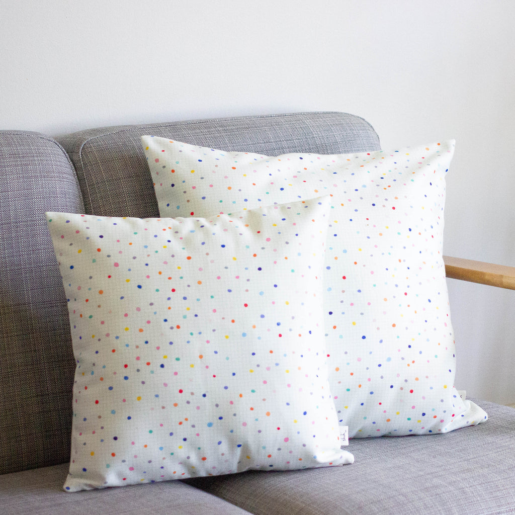 A Front view of the day pop cushions. Two cushions on a couch. Print is multicoloured polka dots on a light grid background. 